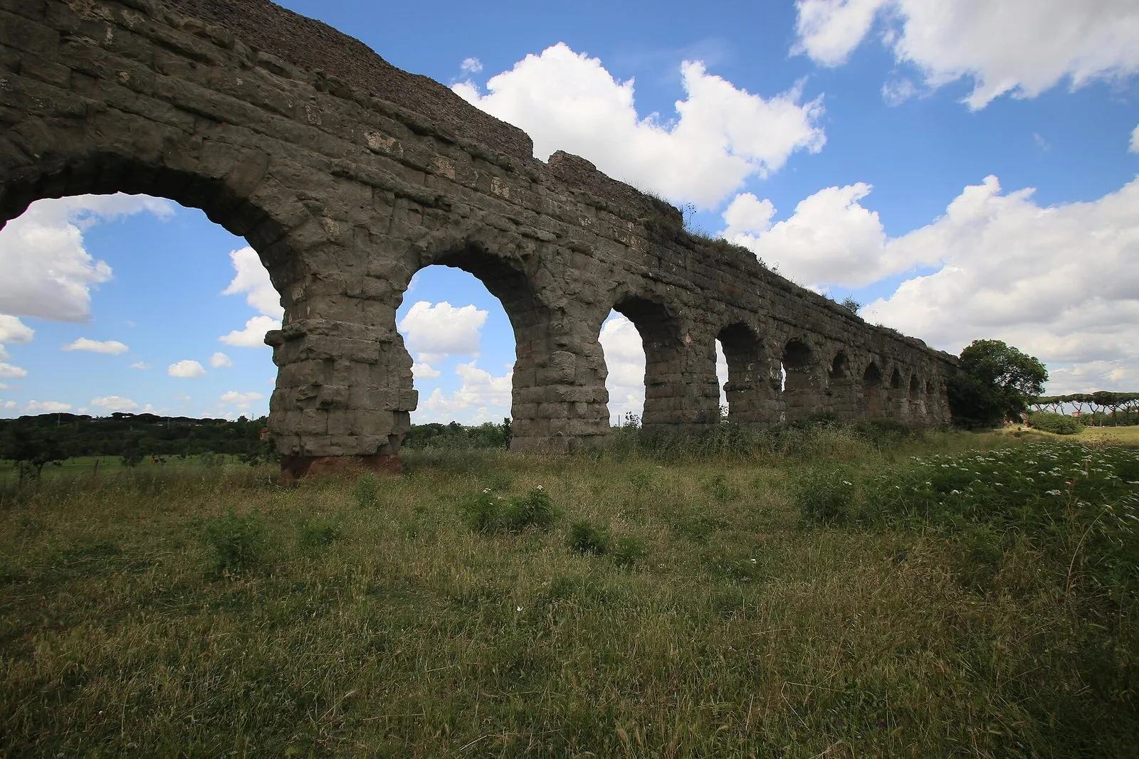 Gli archi in pietra di un acquedotto romano nel Parco degli Acquedotti, tra il prato e un cielo azzurro con nuvole